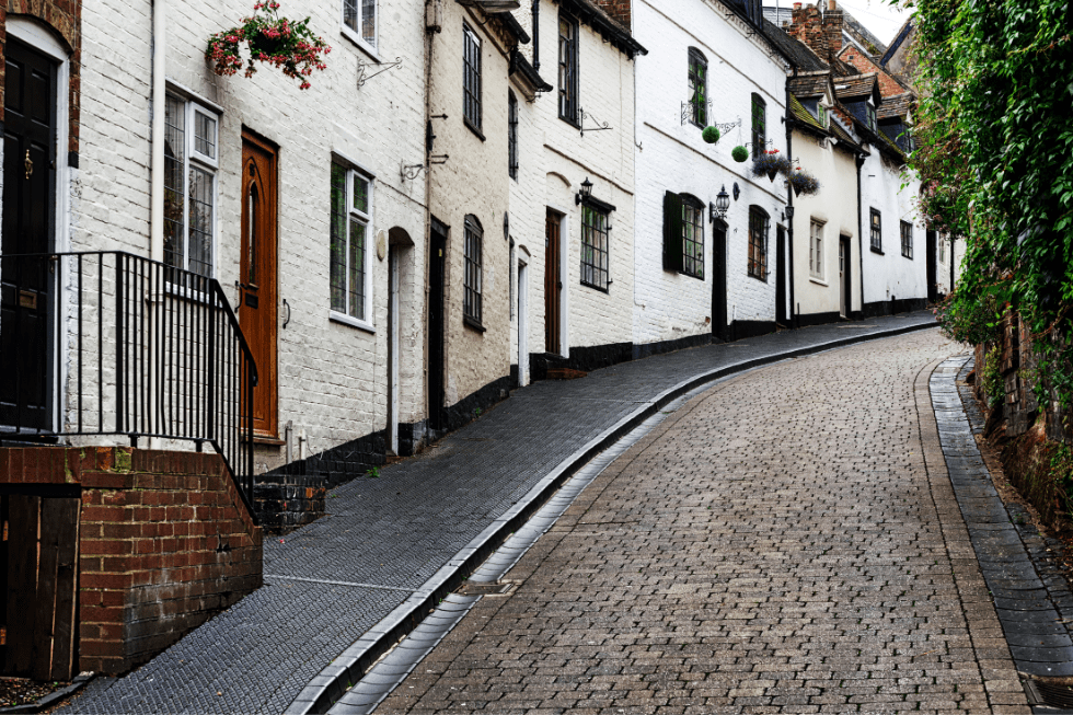 A charming, winding cobblestone street lined with traditional white-washed houses adorned with flowering baskets, following a gentle curve in a historic town.