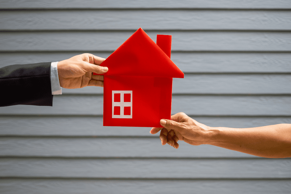 Two hands exchanging a stylized red house cutout against a blurred grey wooden background, symbolising a real estate transaction or ownership transfer.