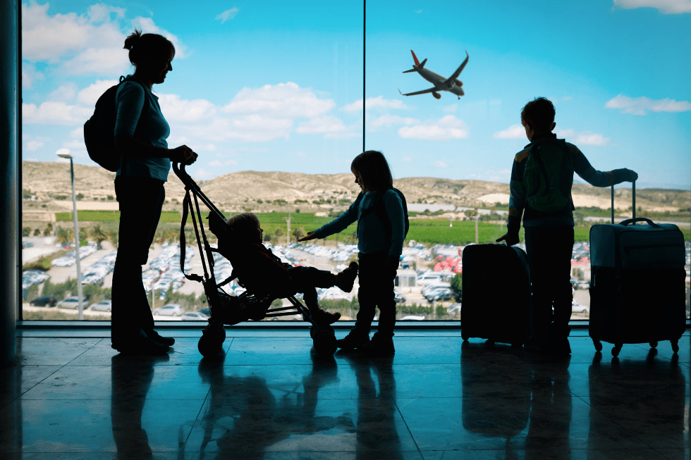 Silhouettes of a family at an airport terminal window watching a plane take off. A woman holds a camera, a child points to the plane, and another child pushes a stroller. Luggage is beside them.