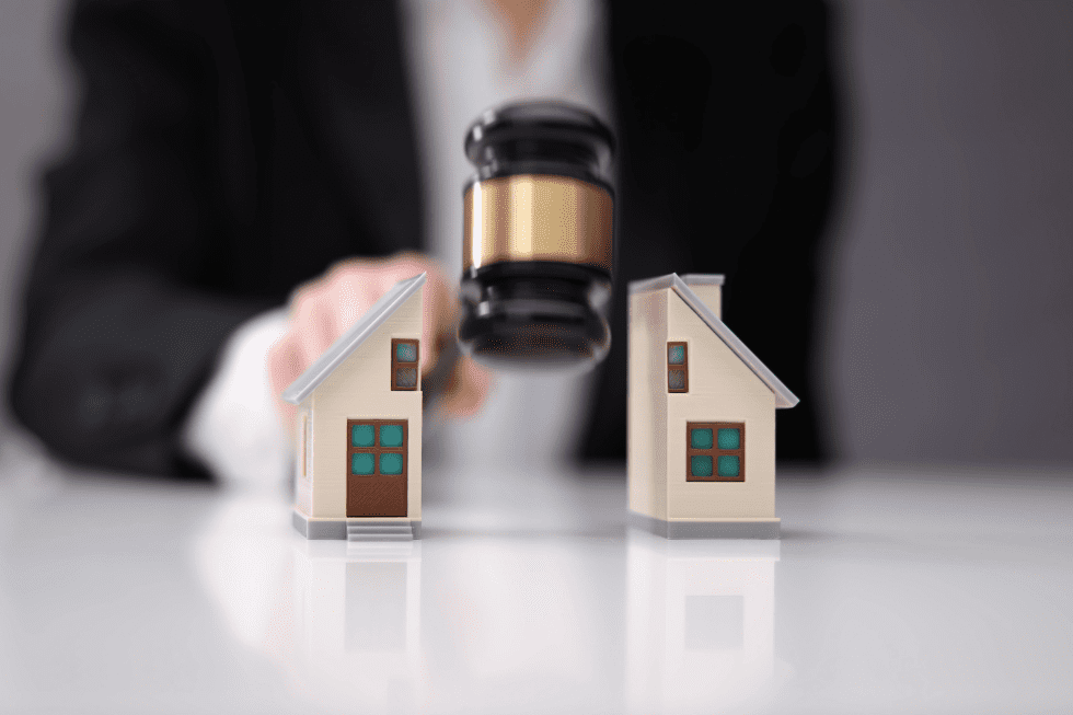 A focused image of a gavel positioned between two model houses, with a person in a business suit sitting at a table blurred in the background, symbolising a property auction or legal proceedings related to real estate.